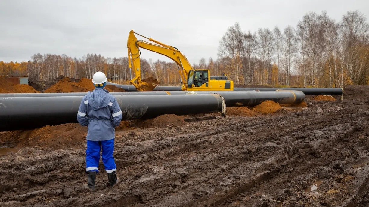 Worker in hard hat stands in front of oil pipeline under construction with yellow heavy equipment.