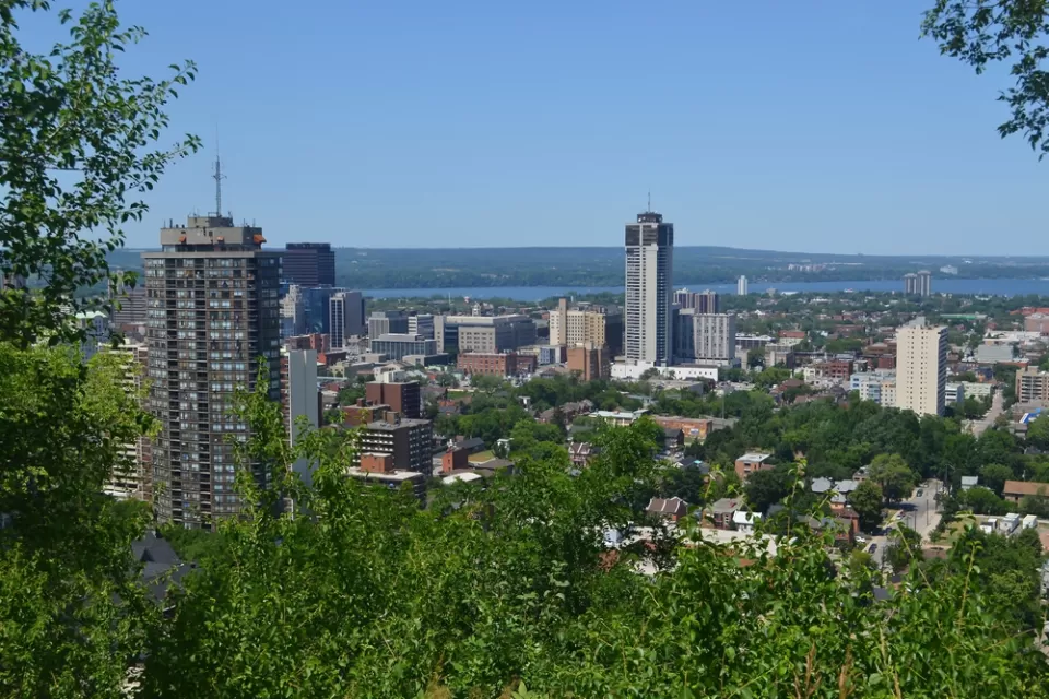 The downtown high rises of Hamilton, Ontario are surrounded by greenery and the blue of Lake Ontario on a sunny day.