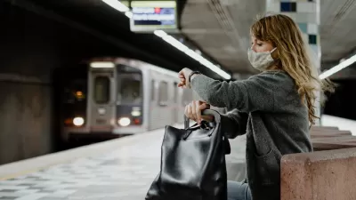 Woman with long hair wearing Covid mask sitting on underground train station bench looking at her watch as subway train approaches in background at Hollywood/Western station in Los Angeles, California.