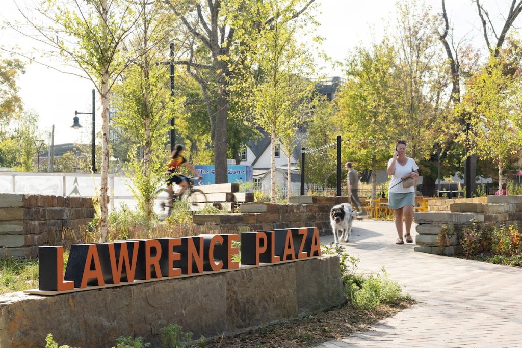 Image of public plaza with large letter 'Lawrence Plaza' sign, person biking by and woman walking in Raleigh, North Carolina.