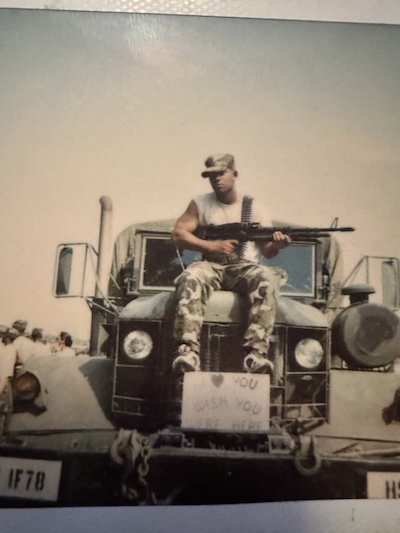 Young Charles holding a firearm sitting on a military Jeep wearing fatigues.