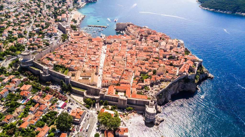 Overhead shot of stone walled city and hundreds of terracota roofs, surrounded on three sides by blue ocean.