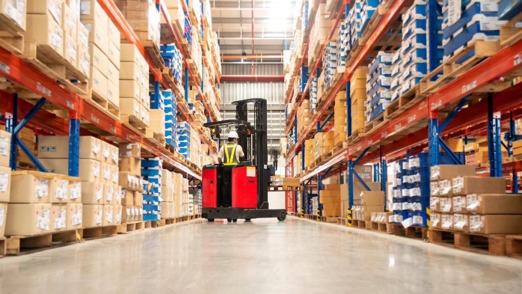 Worker on forklift in large warehouse full of boxes on tall shelves.