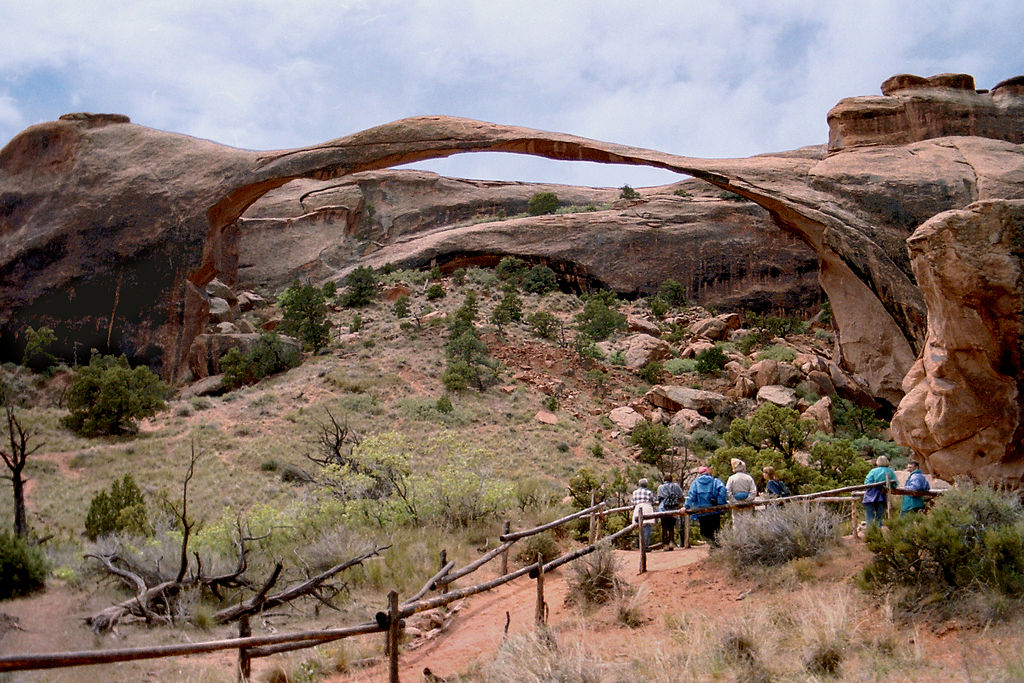 Tourists walk along a red-desert path in Arches Naitonal Park in Utah toward a natural stone arch.