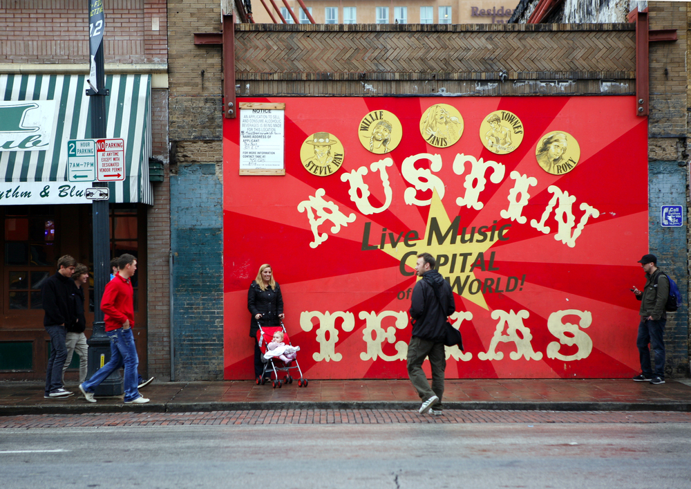 people loiter in front of a large sign that reads, "Austin, Texas: Live Music Capital of the World."