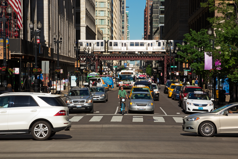 A busy Chicago street, full of cars, pedestrians, and people on bikes. In the background a CTA train passes by on elevated rail tracks.