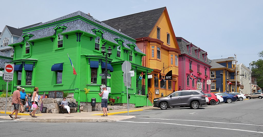 A row of colorful houses, some with businesses on the ground floor. Numerous people are standing and walking on the street.