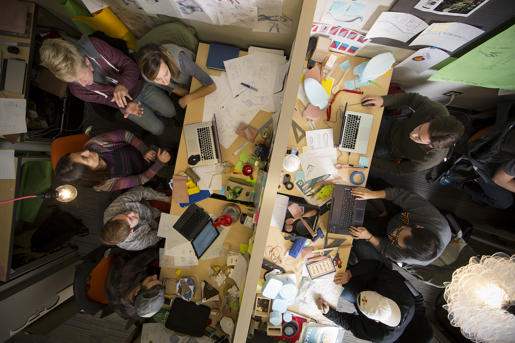 Image of areial view of group of students around a square desk in small room full of papers, computers, and books