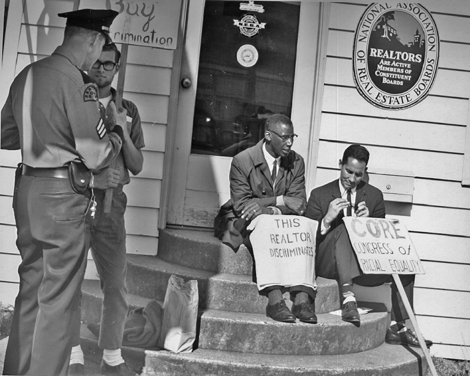 Two men sitting on steps of realtor office in Seattle in 1964 holding protest signs that accuse the realtor of discrimination.