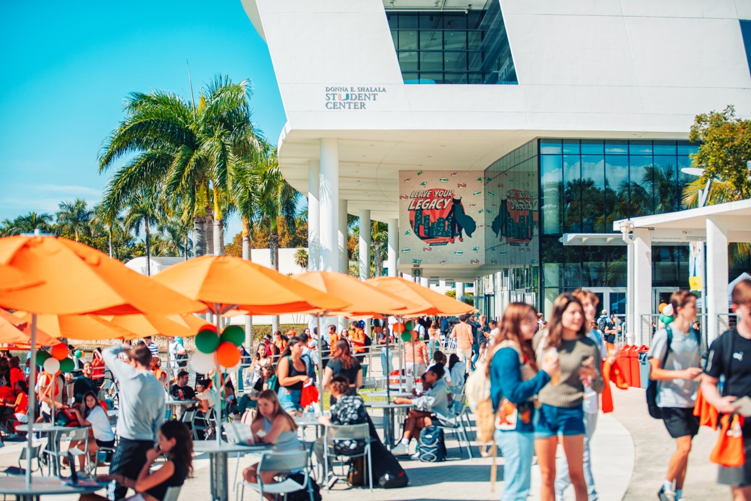 Image of a busy food court like area with round tables with orange umbrellas with palm trees in the background