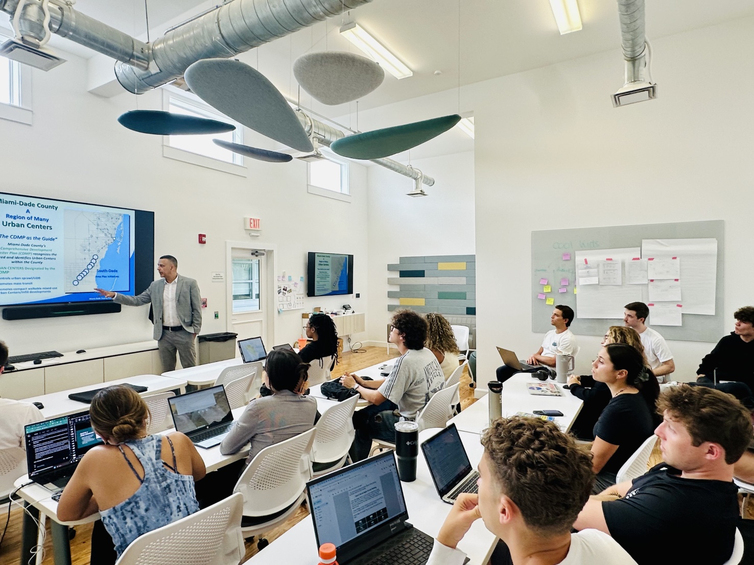 Classroom full of students with open laptops listening to the profressor in the front pointing at a map