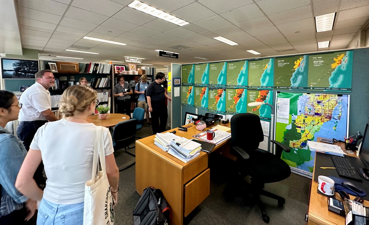 Image of an office room filled with people standing who are looking at a wall with maps plaster on them multiple times with varying differences