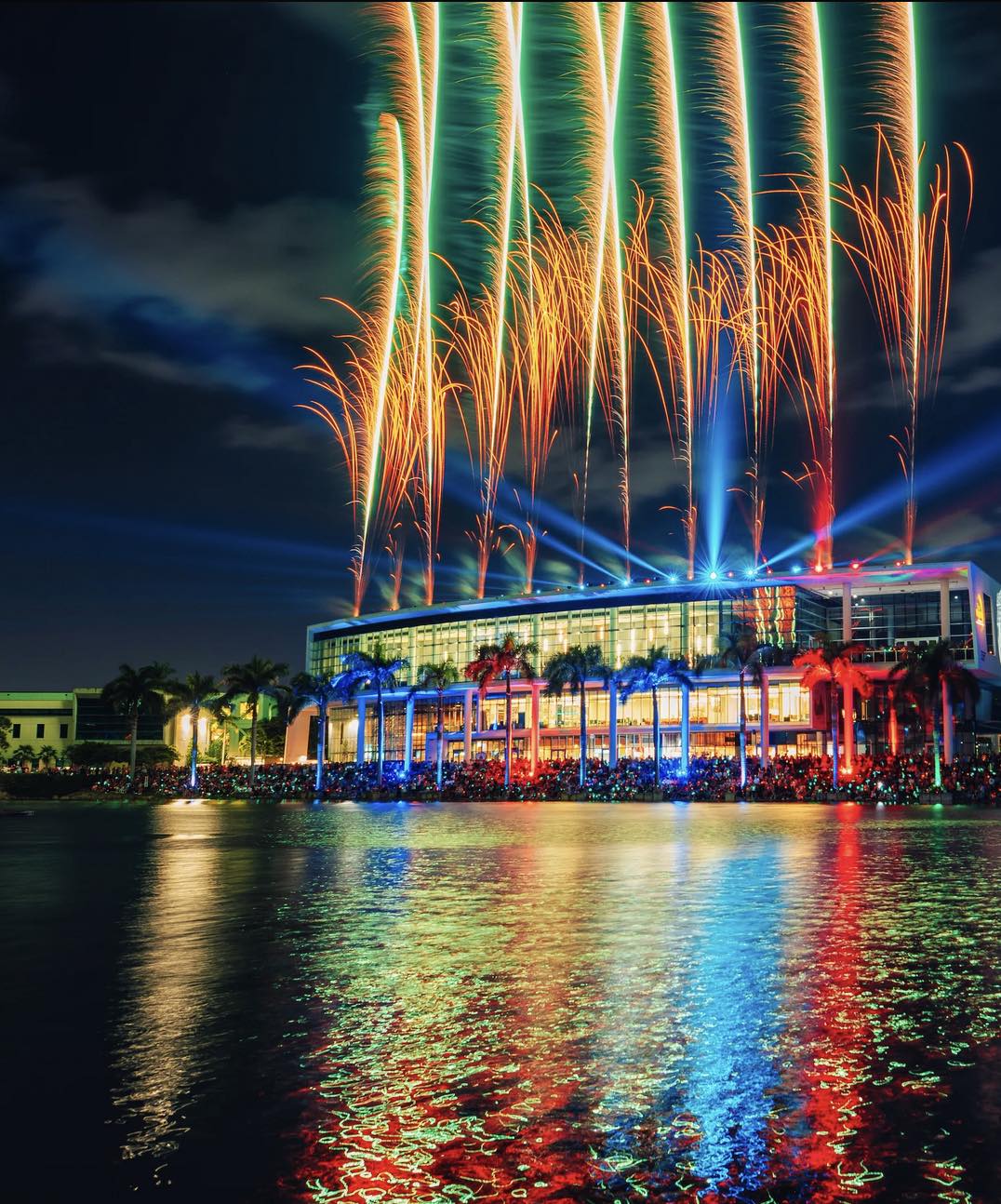 Image taken at night of a building sitting next to water with fireworks light across said building with the reflections of the colors bright against the water