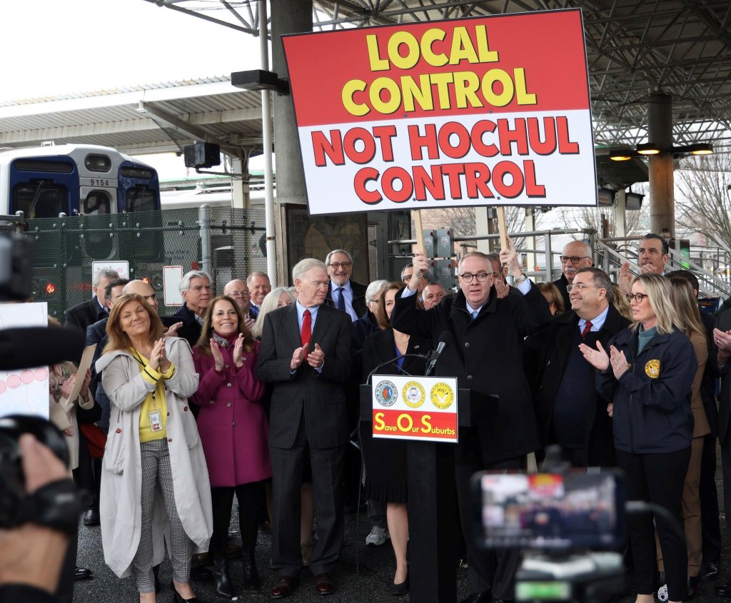 Hempstead Town Supervisor Don Clavin holds sign which says "Local Control not Hochul Control" at a podium while standing with many other local elected officials at a train station.