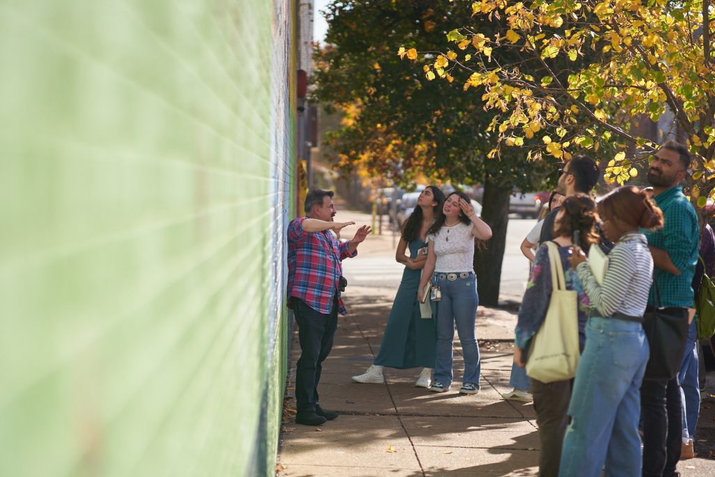 Image of an instructor speaking with his students outside against a green wall with trees in the background