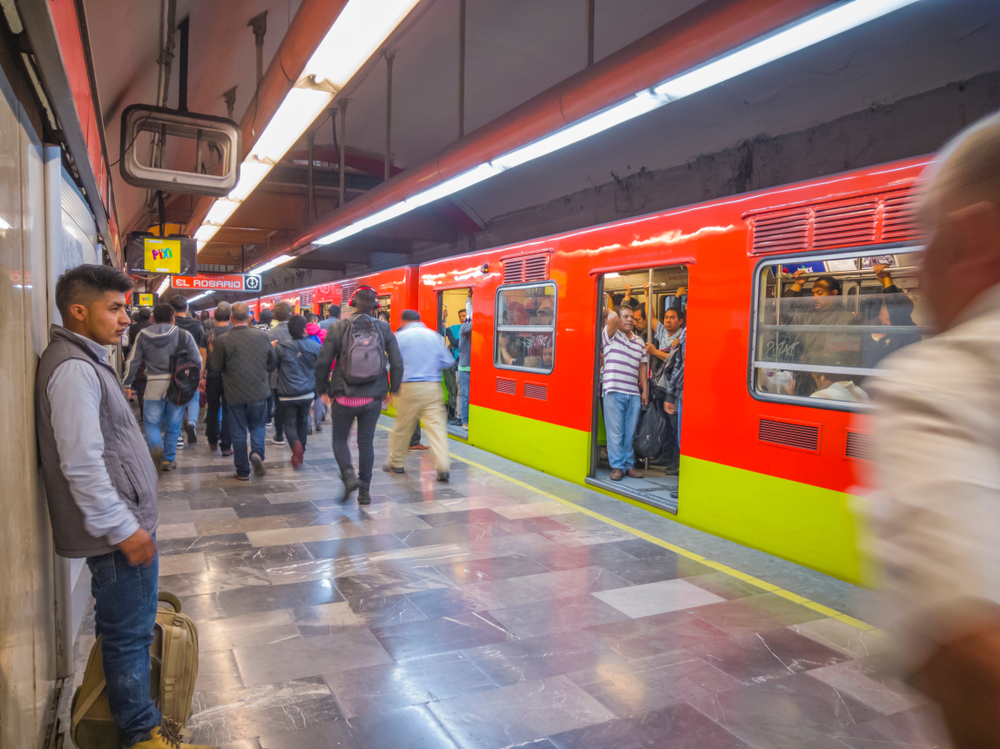 A passenger waits for a train in the Mexico City Subway, while passengers board and alight a train in the background.