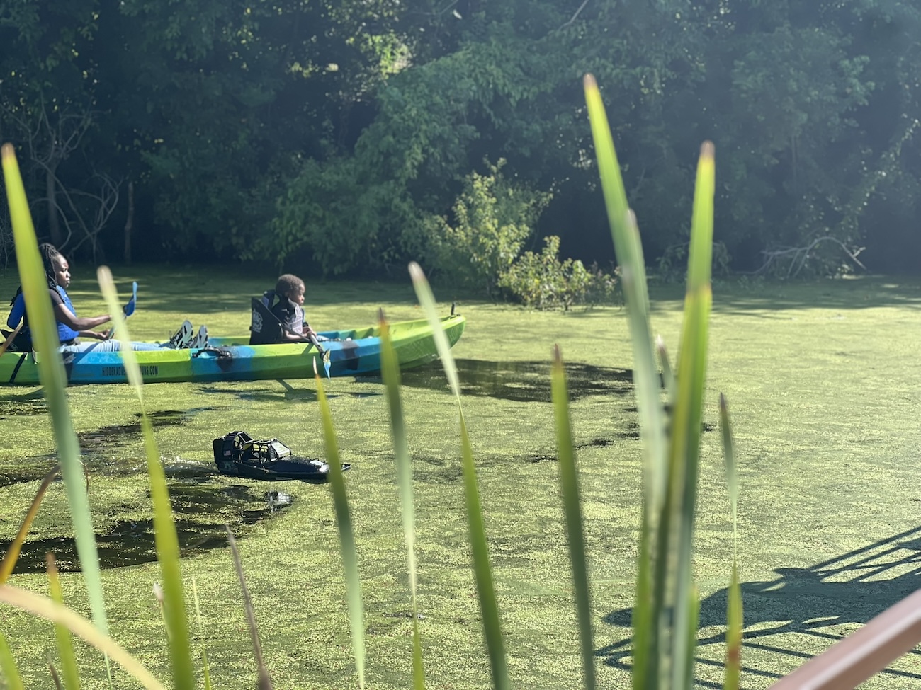 A mom and son kayak near a remote control airboat in Sankofa Wetland Park. Image: Sankofa Community Development Corporation. 