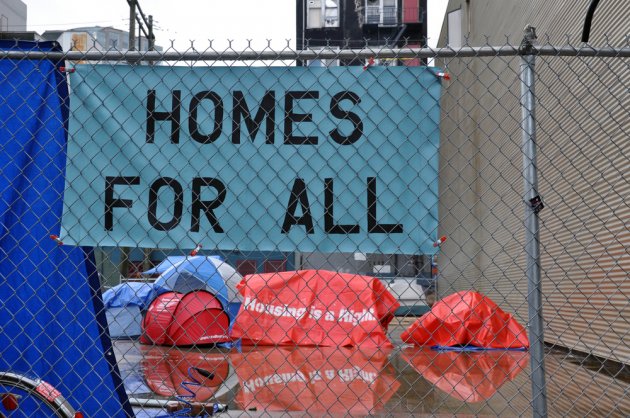 Tents housing homeless people are shown behind a chain link fence and below a sign that reads, "Homes for All."