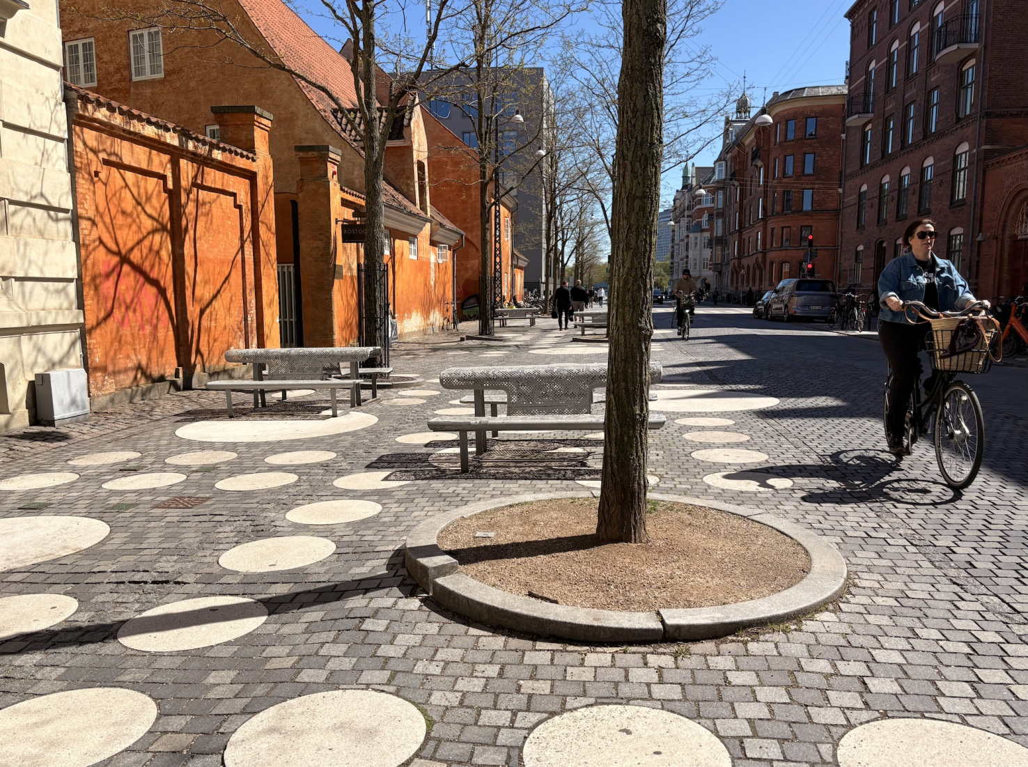 People ride bikes past pedestrian plaza with white circles in paving stones next to old buildings in Copenhagen, Denmark.