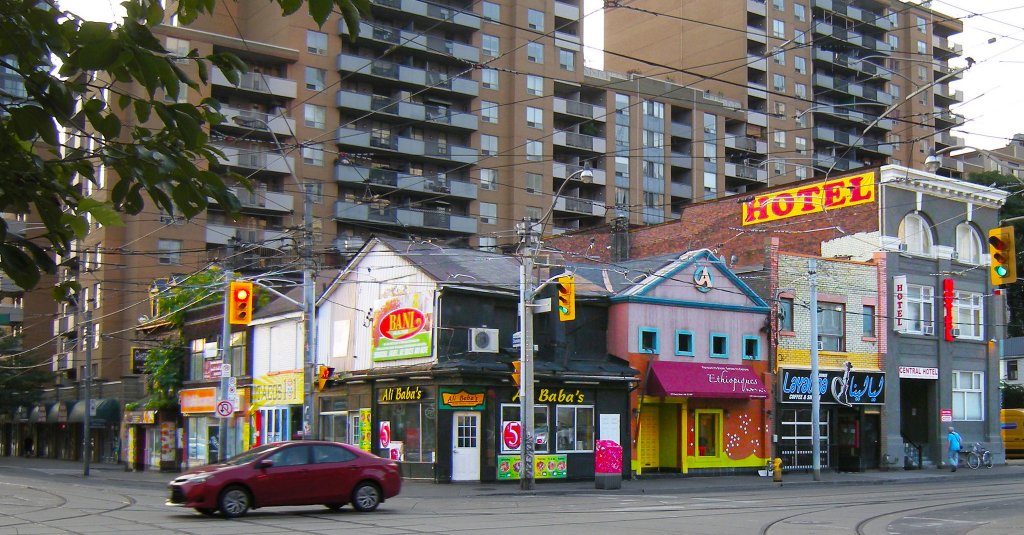 Small but colorful shops with second-floor apartments dot a street corner, while high-rise apartment buildings loom in the background.