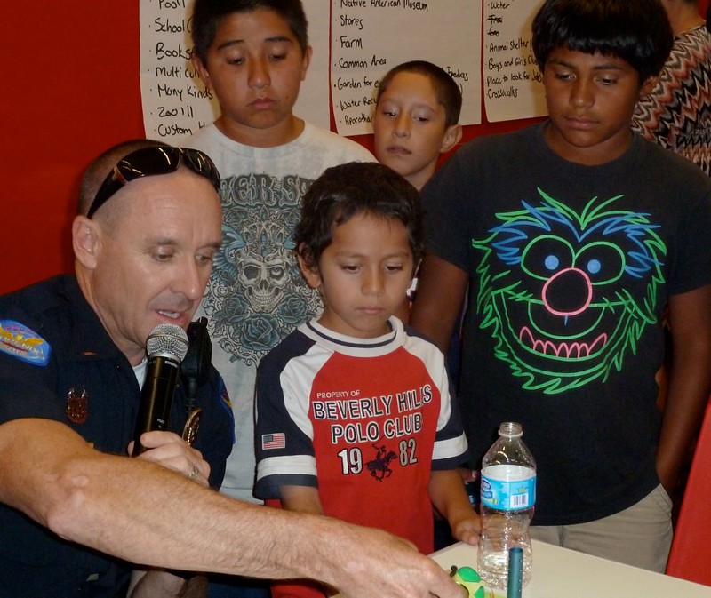 A small group, including a police officer in uniform and several small children, discusses maps they made from toys.