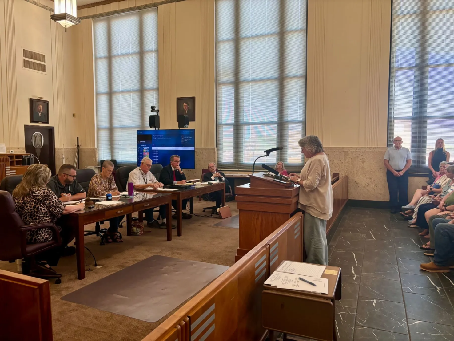 A woman with gray hair stands behind a podium and speaks to a six-person board in a while room with tiled floor.