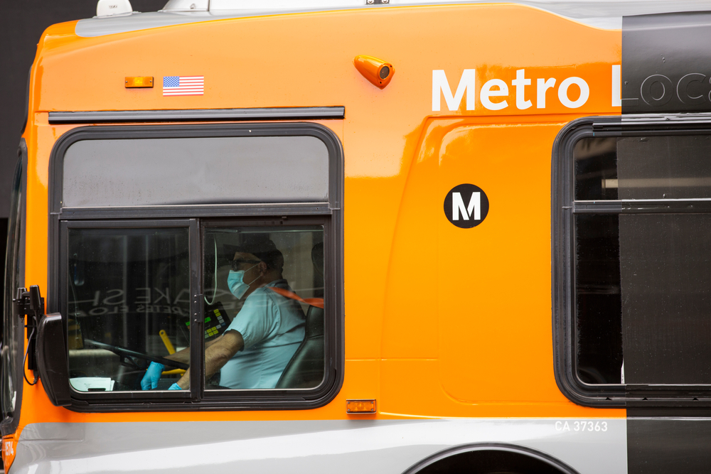A bus driber wearing a mask turns the wheel of a large orange public transit bus.