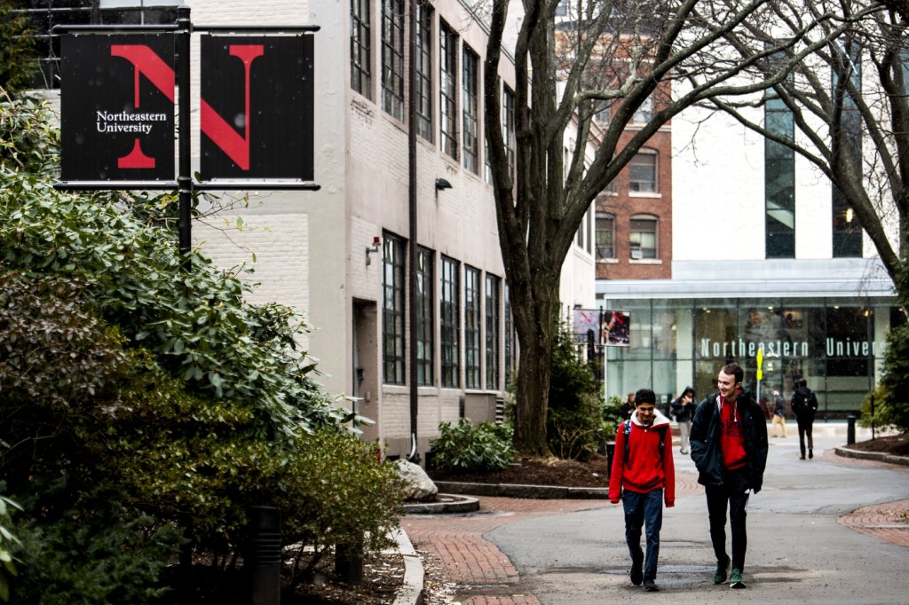 Image of a pathway between buildings on the Northeastern University campus with two male students walking and chatting with each other on a brick and cement pathway.