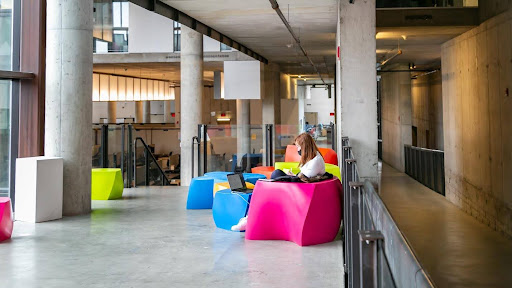 Image of a student studying on a geometric shaped pink chair with other geometric shaped furniture surrounding them. The colors being yello, pink, and blue within a wooden mixed with concrete building with concrete pillers