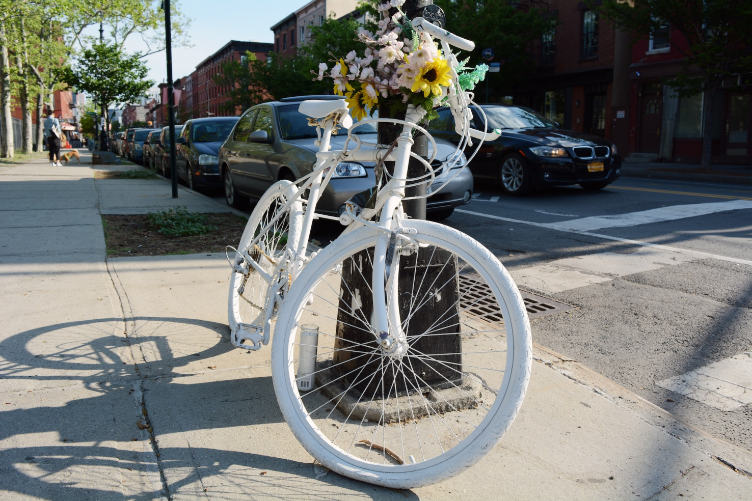 White "ghost bike" with pink and yellow flowers tied to a black light post on sidewalk.