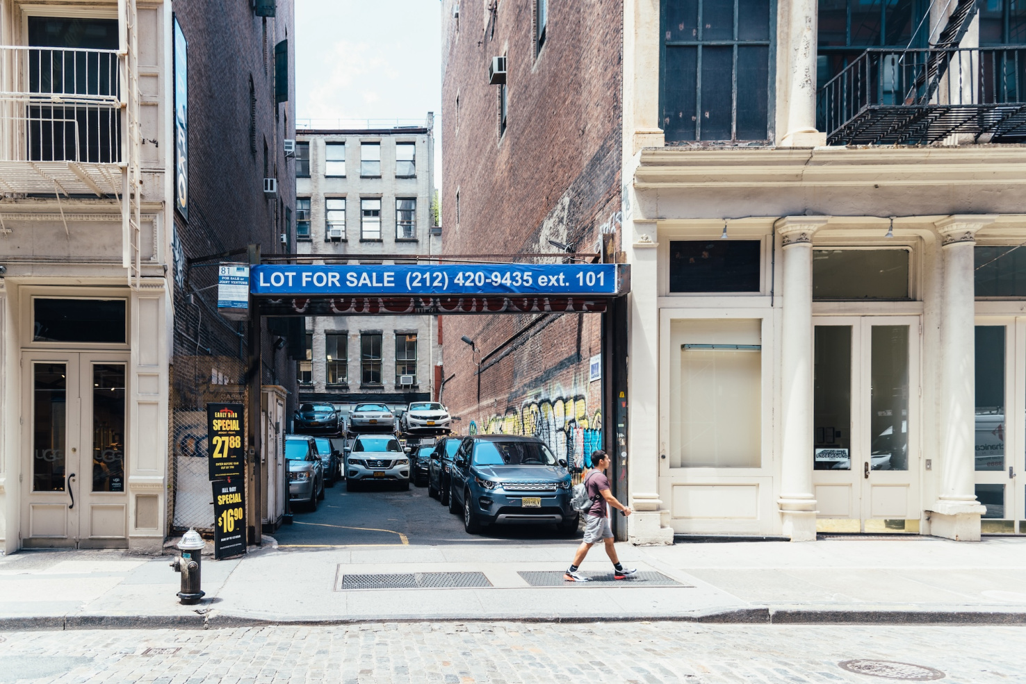 Parking lot with "Lot for Sale" sign with phone number on blue banner between brick buildings in New York City.