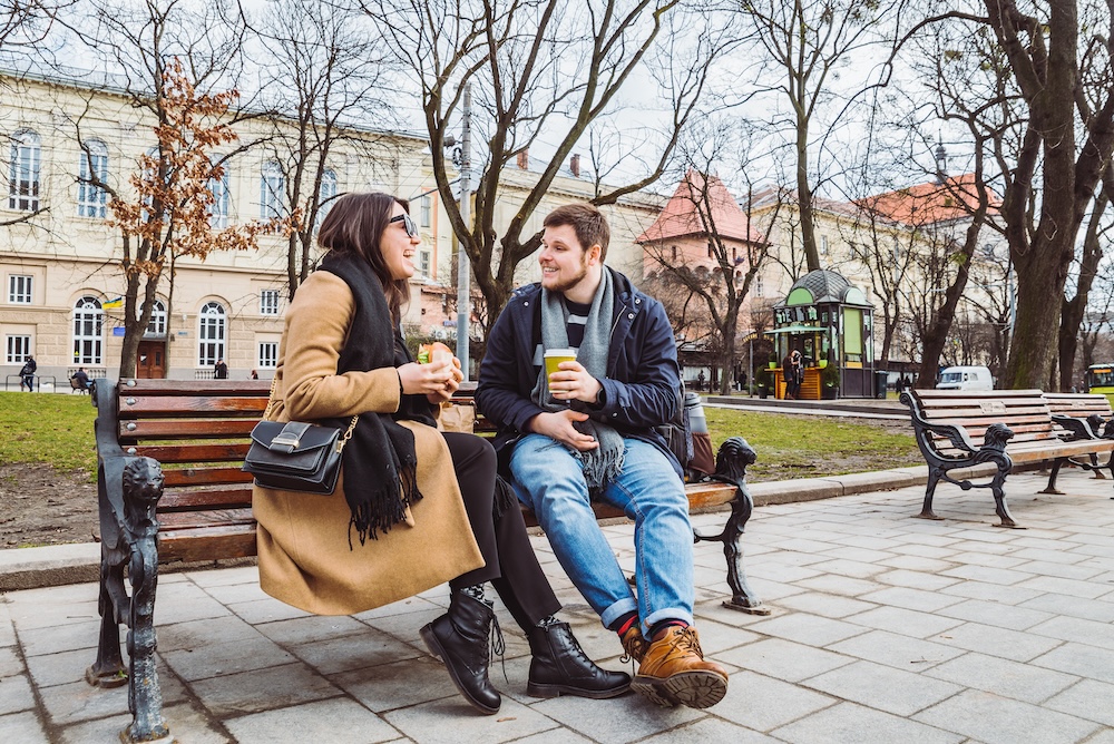 young adult couple eating fast food on bench of city park.