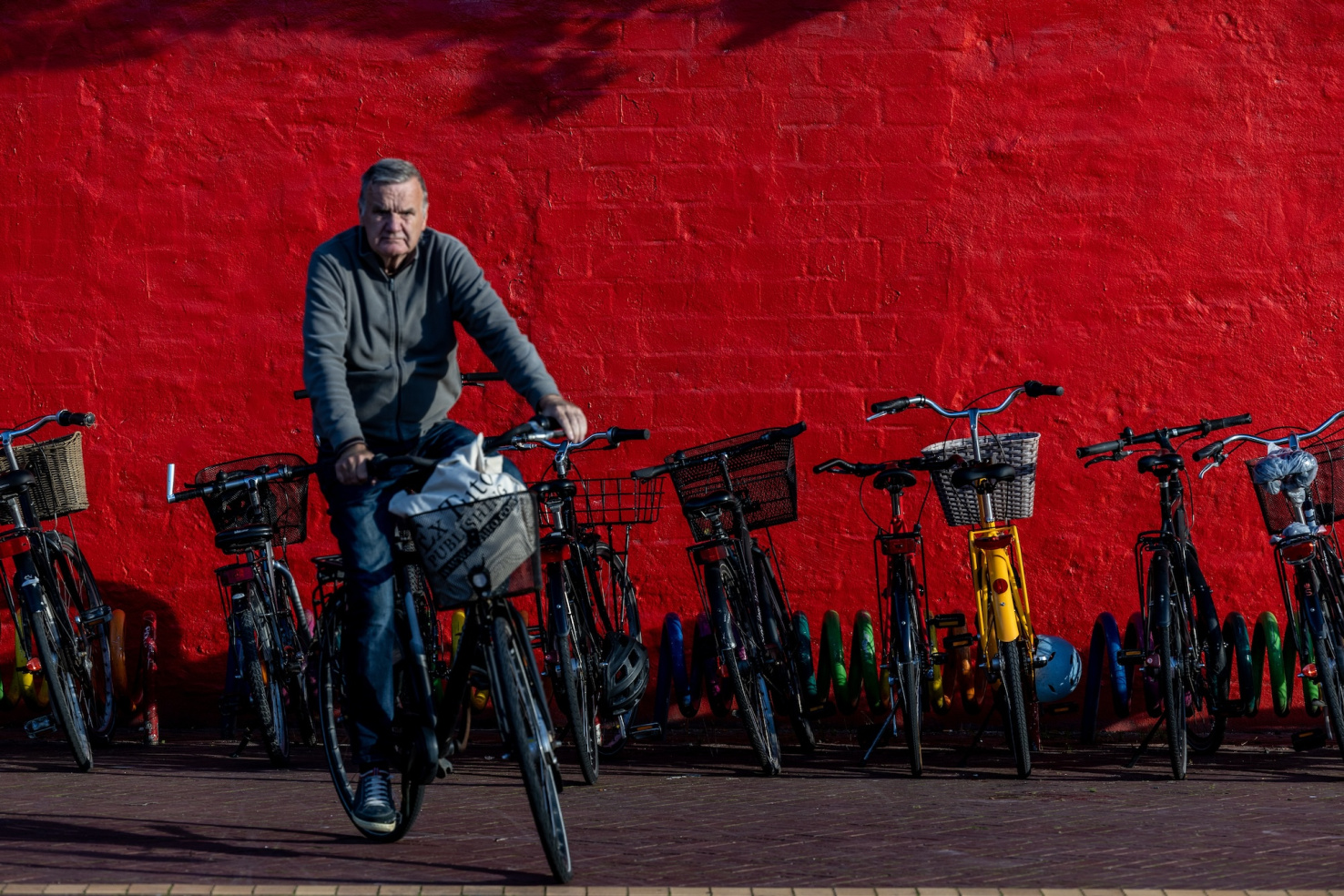  A senior man bicycles past a row of bikes along a red wall.