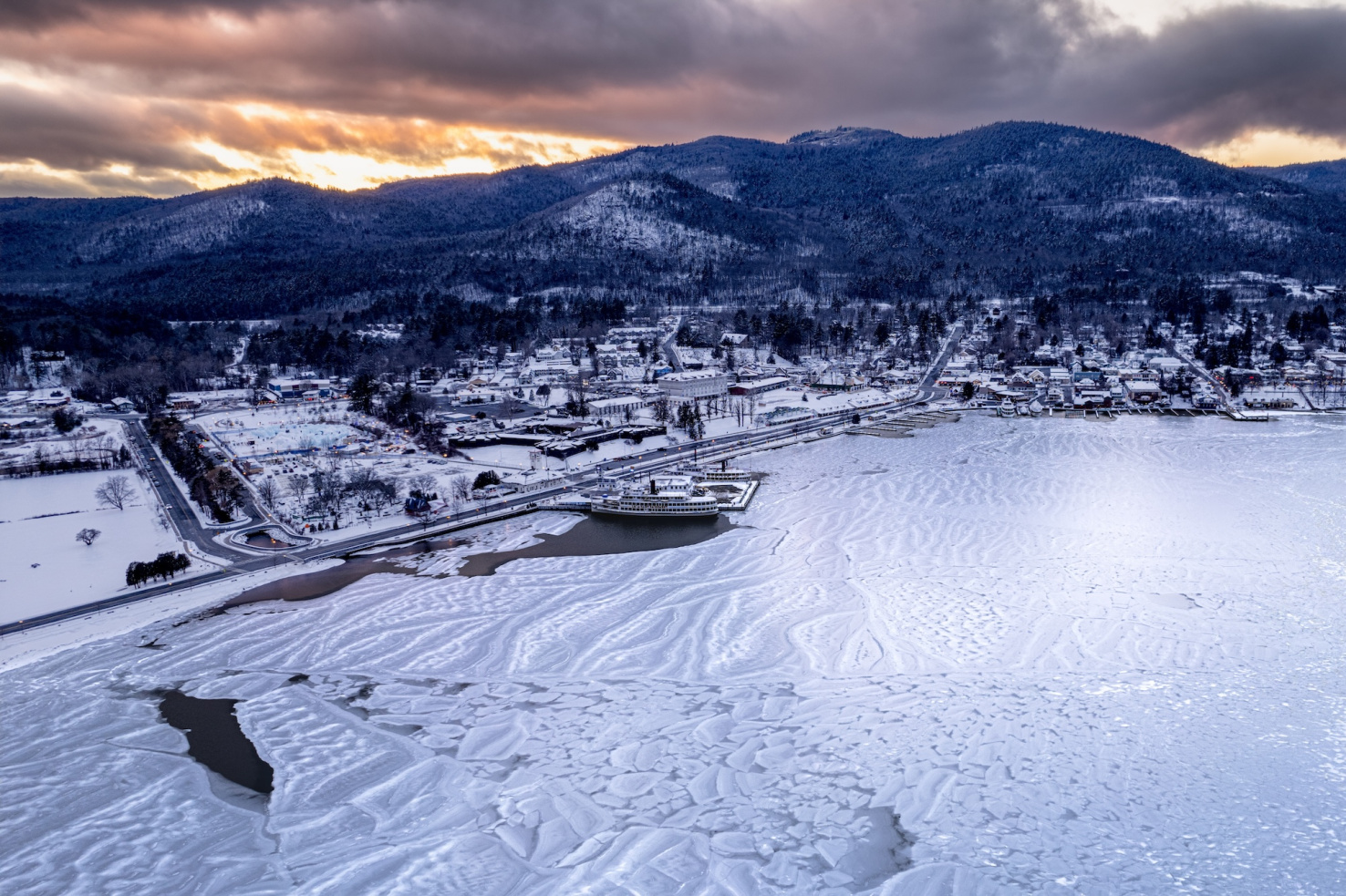 Aerial of frozen Lake George, New York at dusk with snowy hills behind the lake.