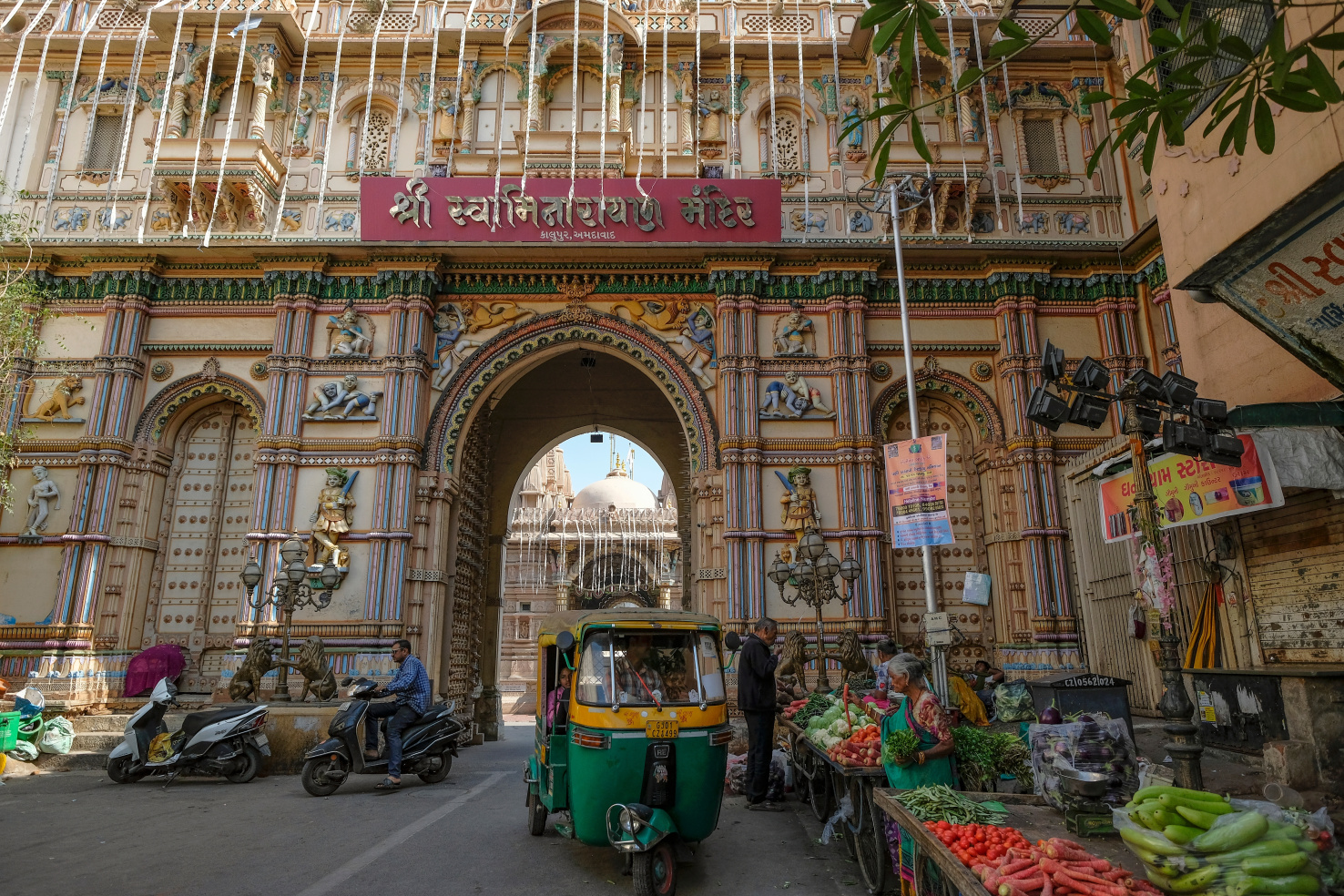 People sell various vegetables in front of a temple, which is ornately decorated. Two mopeds are also visible.