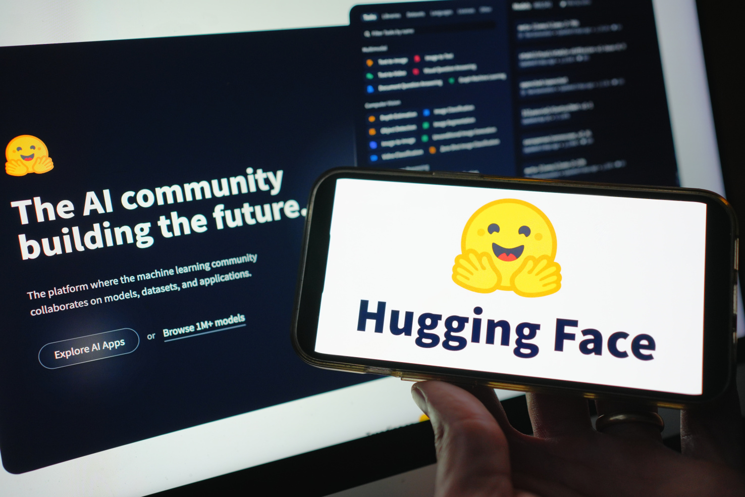 The Hugging Face logo, a smiling yellow face, with the text "The AI community building the future" one a computer screen in the background.