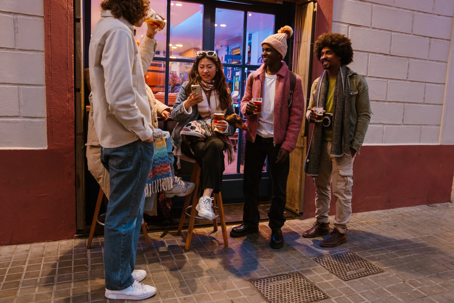 Group of friends standing and sitting on a stool outside a bar or cafe at night.