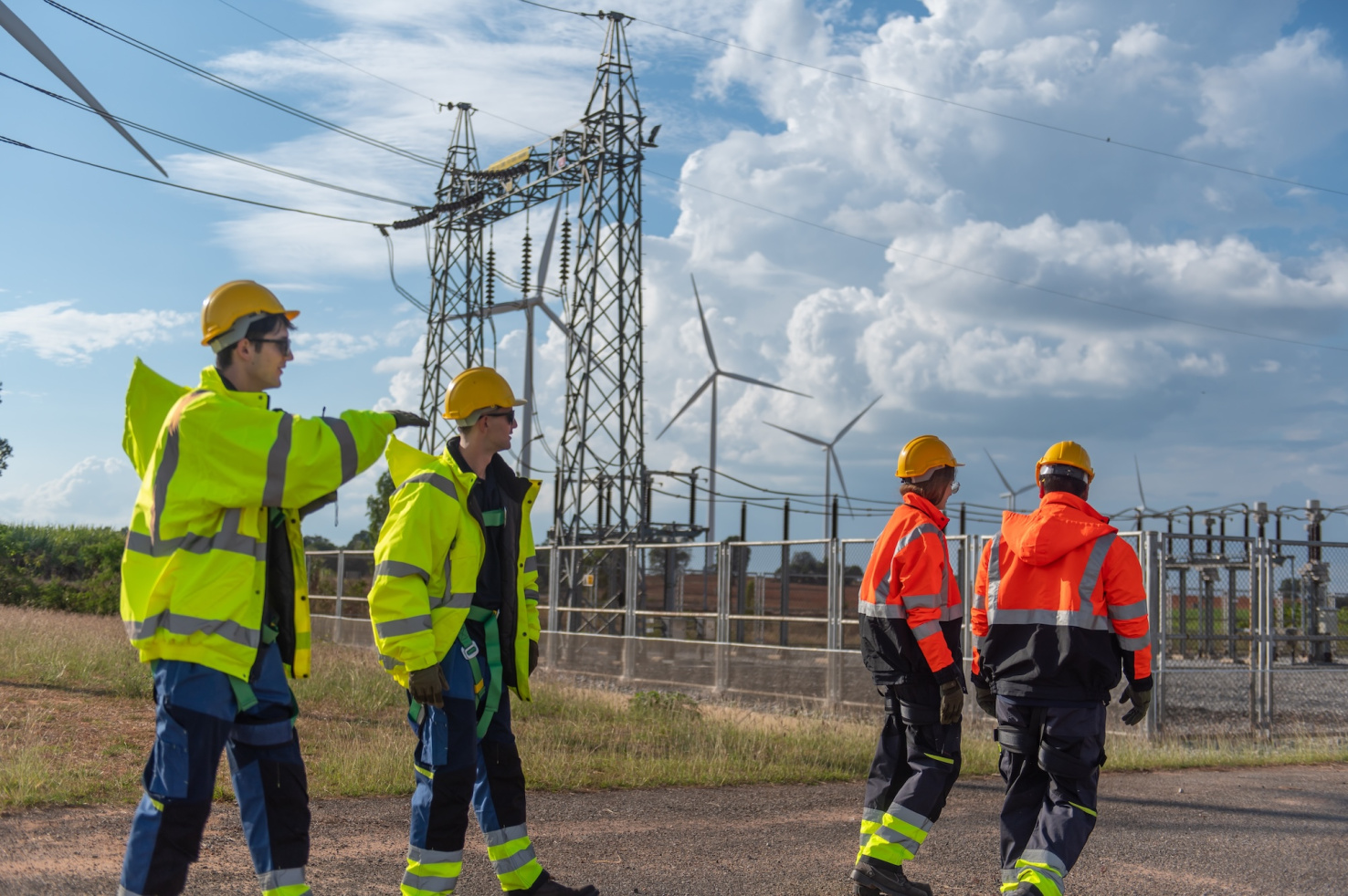 Workers in yellow and orange safety jackets walking next to power transmission lines and wind turbines in a field.