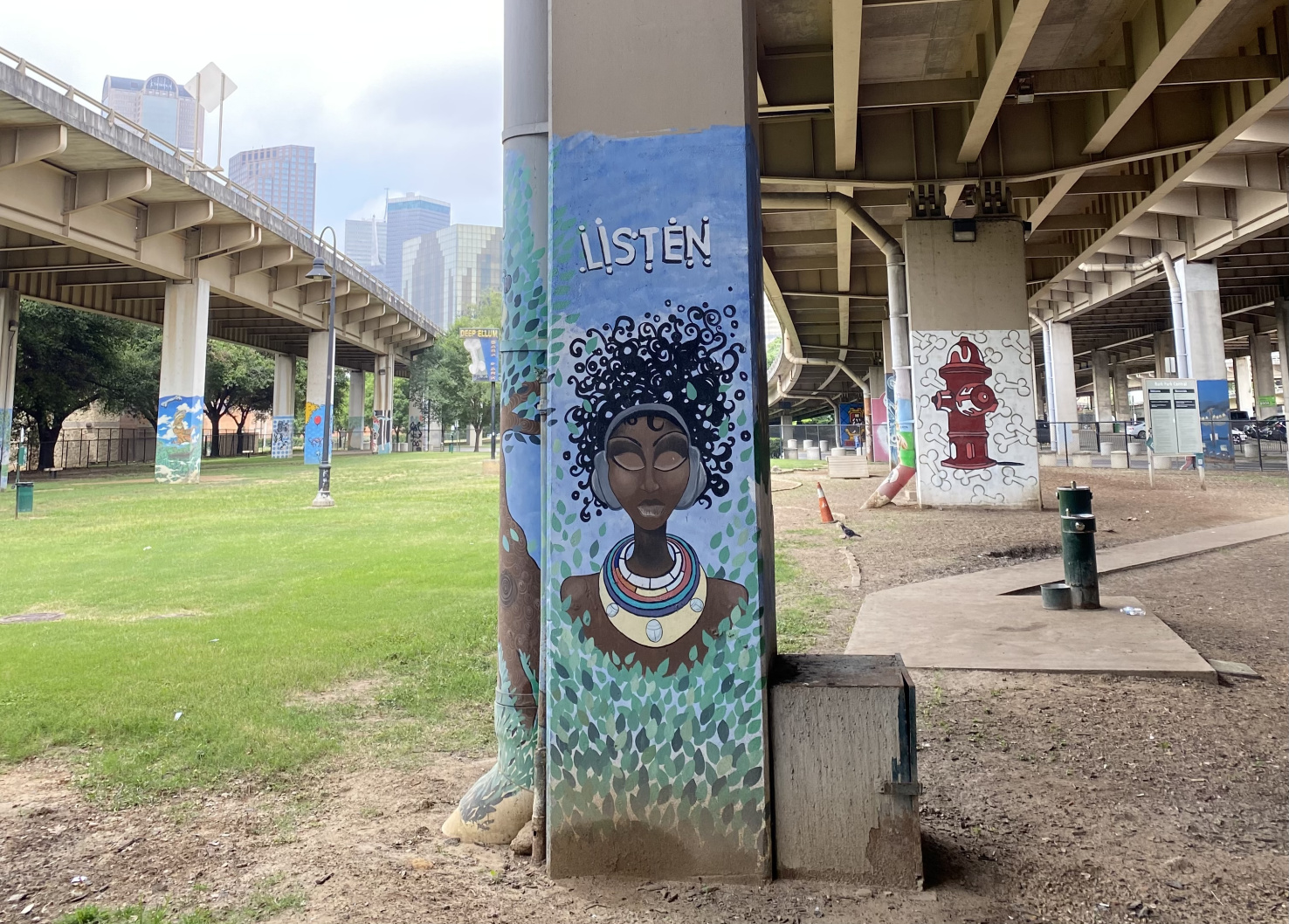 Under a highway, support beams are painted with pictures. One has a fire hydrant and dog bones, another has a Black woman with headphones and the word 'listen' written above her