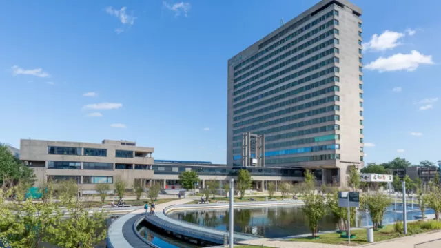 Image of a tall long rectangular building next to a large body of water with walkways and a bright blue sky with some clouds