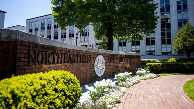 Image of the brick sign of Northeastern University's sign and logo with a small garden and brick pathway in front of it and white buildings in the background