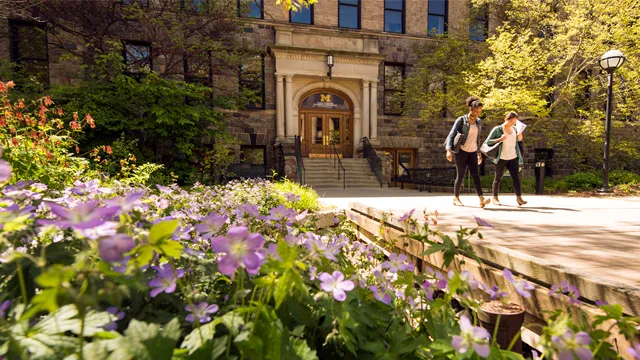 Image of two students walking away from the University of Michigan Dana building at ground level with a bed of flowers framing the photo