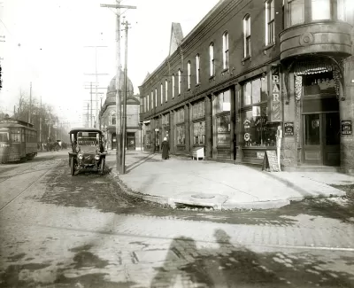 Black-and-white photo of street with old black model T and brick building on the corner.