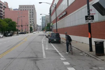 Man on bike looking back at camera with black car blocking bike lane in front of him.