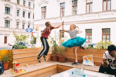 Parklet in Vienna with two young girls jumping up to high-five each other, one wearing a bright teal tutu.