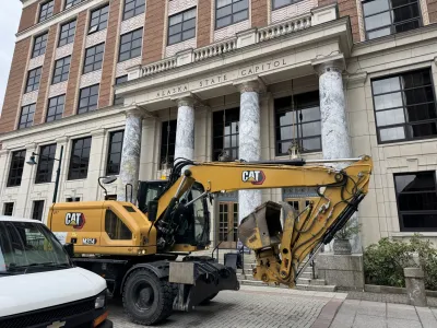 A CAT excavator drives past a brick building with marble columns.