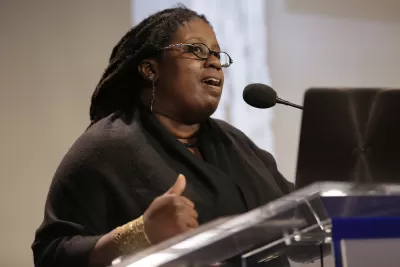 Black woman speaking at podium with presentation screen in background.