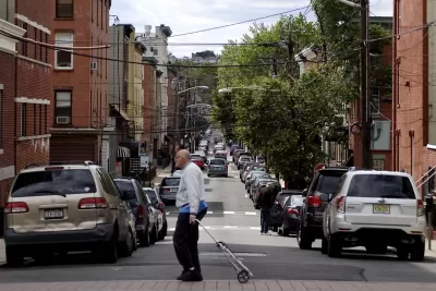 An old man crosses a one-way street in the foreground, with both sides of the streets lined with cars facing away from the camera and brick buildings.