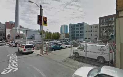 A surface parking lot, full of cars, is located between tall buildings and urban density.