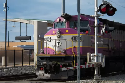The front of a purple and gray train running along track with a rail crossing signal on the right.
